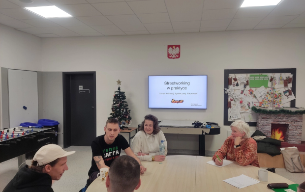 Grupa ludzi zasiada wok&oacute;ł stolu i&nbsp;rozmiawiają. W tle tablica interaktywna z&nbsp;tematem rozm&oacute;w "Streetworking w&nbsp;praktyce".